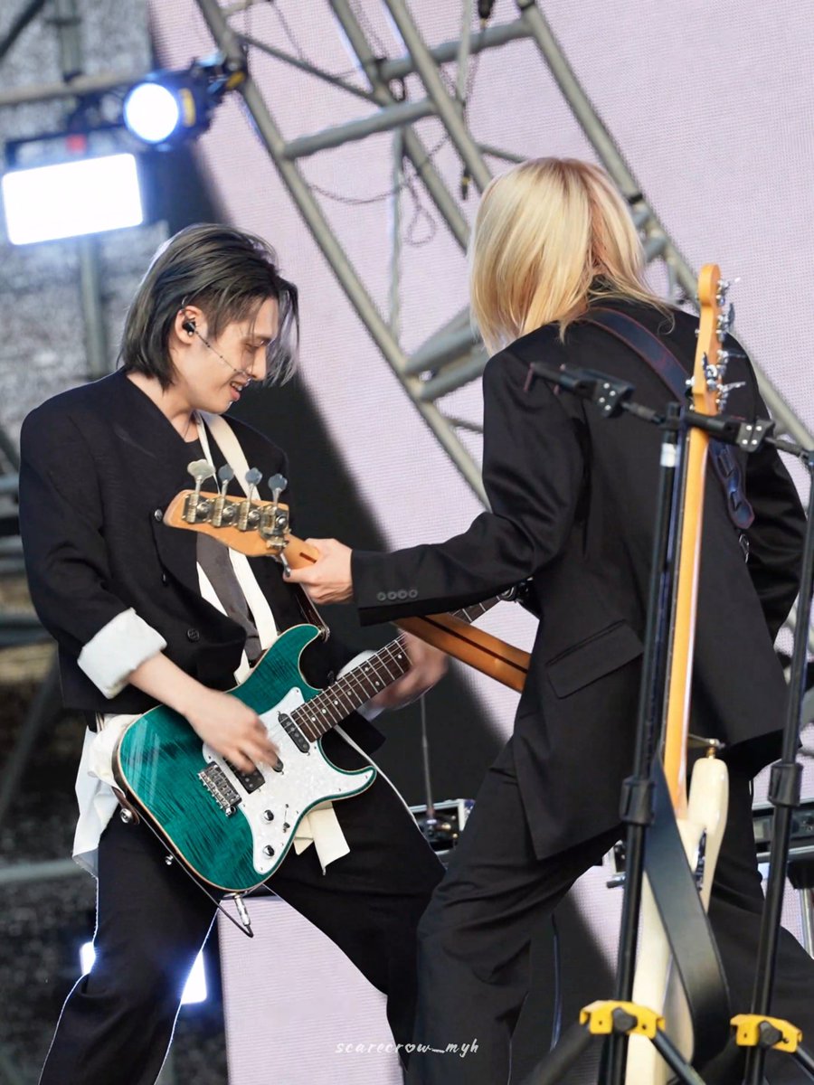 Two musicians in matching black suits perform energetically on stage, with the bassist playing a striking teal bass guitar as the blonde guitarist faces away, sharing a moment under bright stage lights. The watermark "scarecrow__myh" appears at the bottom, capturing what appears to be a lively concert moment aligned with the enthusiastic post celebrating performers like Gaon and Juyeon from Xdinary Heroes.