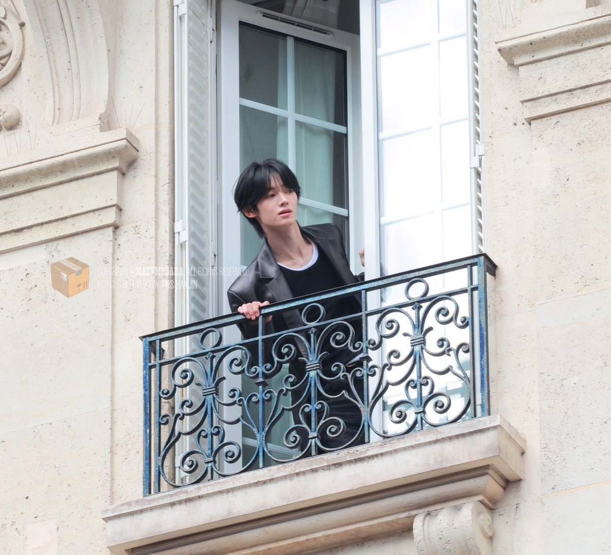 A person stands by an open window behind an ornate iron balcony railing of a grand stone building, leaning casually on the railing in a black blazer and t-shirt. The scene evokes a modern, brooding "trapped prince" aesthetic, fitting the caption's playful comparison to a Disney-style tale of a secluded royal in a castle.