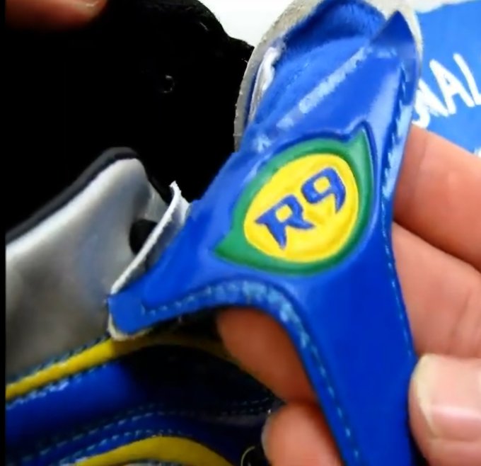 A close-up of a blue and yellow soccer cleat displaying a green and yellow "R9" logo on the tongue, referencing the Brazilian soccer legend Ronaldo.