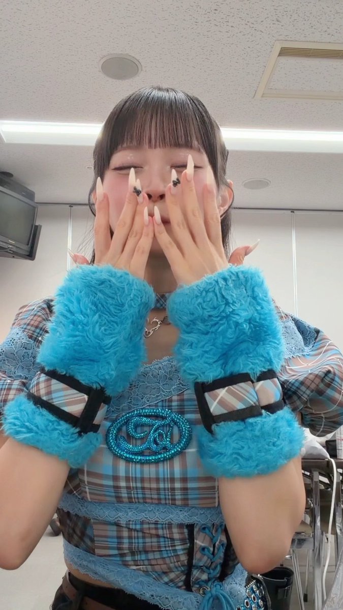 A young woman with bangs covers her face with her long-nailed hands, wearing a blue plaid outfit with fuzzy cyan cuffs and a rhinestone "Z" emblem. Her closed-eye, smiling expression conveys a sense of shy, secretive delight—mirroring the post's theme of something deliciously irresistible that subtly "seeps in."