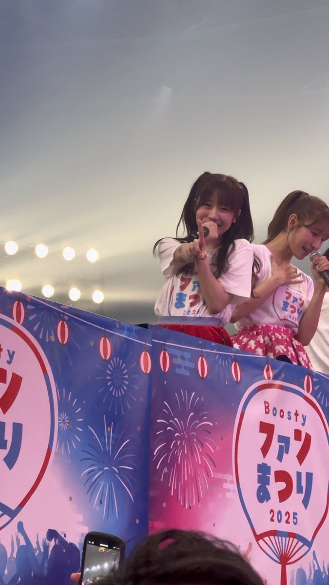 Two young female performers on stage at the 'Boosty Fan Matsuri 2025' event, with one pointing and smiling at the camera while wearing a white t-shirt with the event logo.