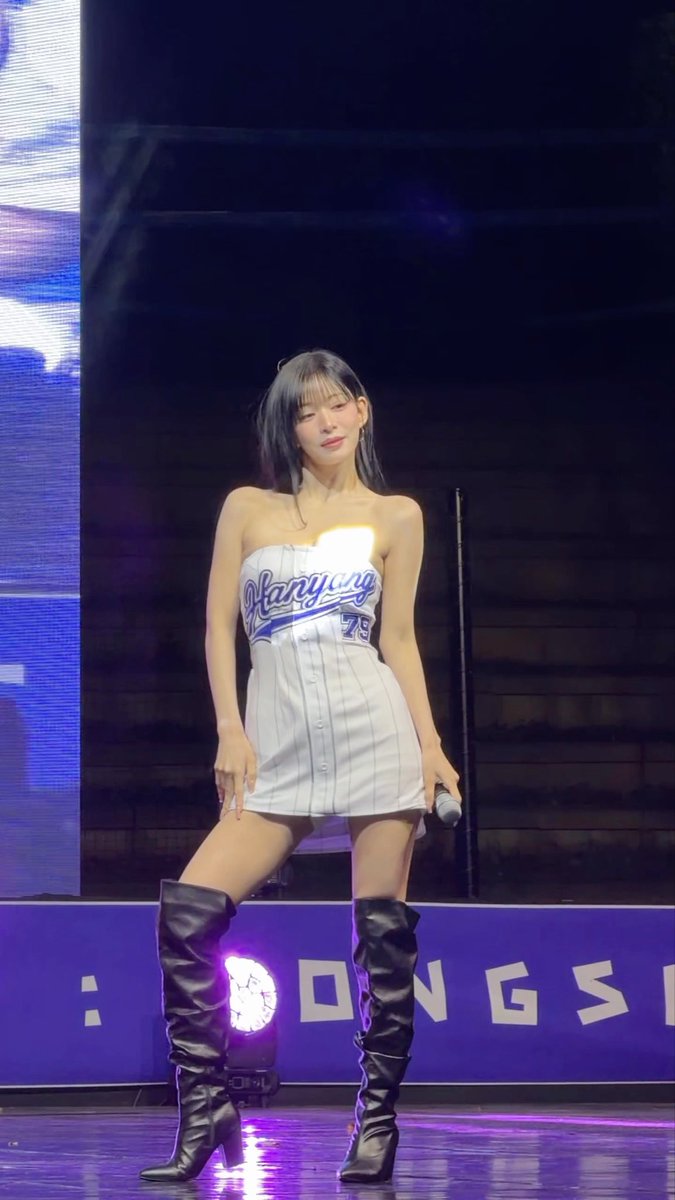 A performer on stage wears a white strapless baseball-style mini dress with blue 'Hangang 79' lettering, paired with black knee-high boots, holding a microphone.