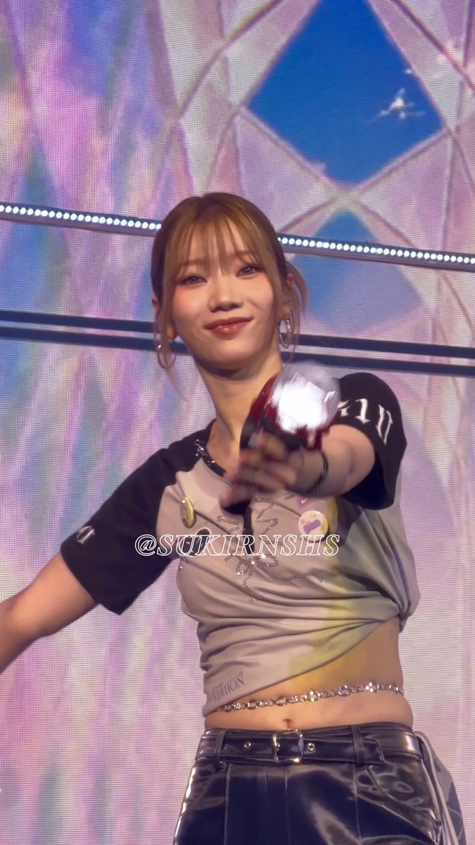A young woman with light brown hair smiles on stage, wearing a gray cropped top and black pants, with the watermark '@SUKIRNSHS' visible.