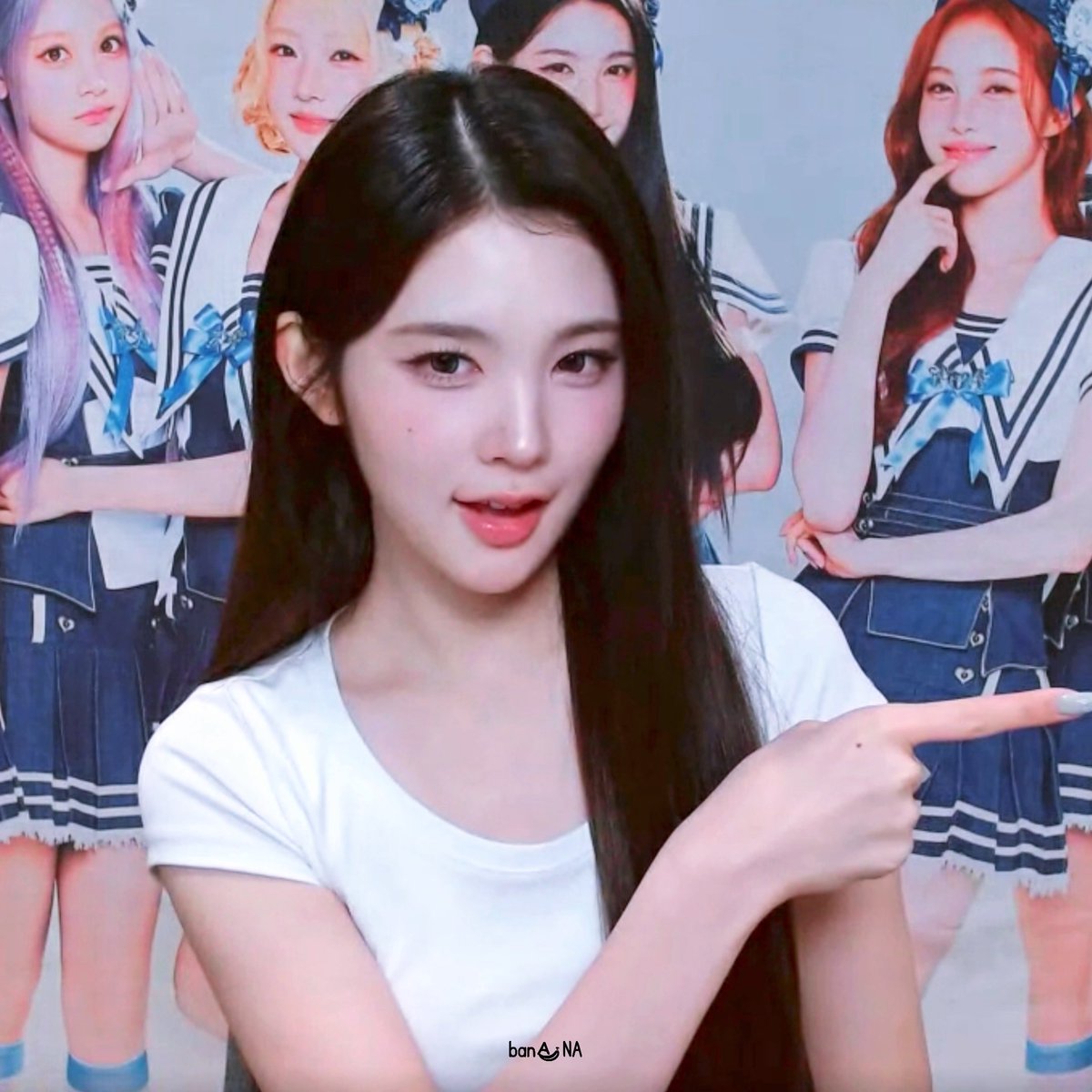 A young woman with long dark hair wearing a white t-shirt points to the right, with K-pop group members in matching outfits visible in the background and a 'banana' watermark below.