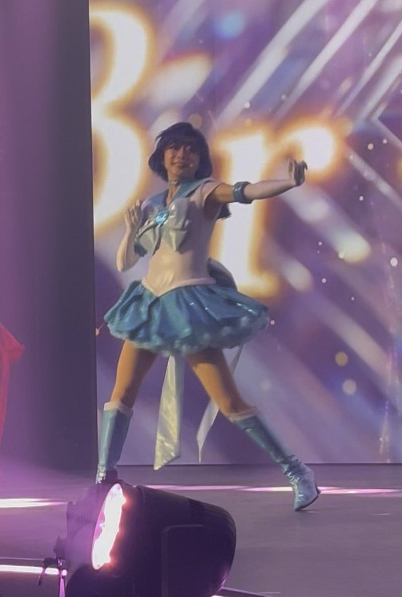 Cosplayer or performer dressed as Sailor Mercury (Sailor Moon character) in a blue and white sailor outfit with knee-high boots, posing on stage with one arm extended against a sparkling purple backdrop.
