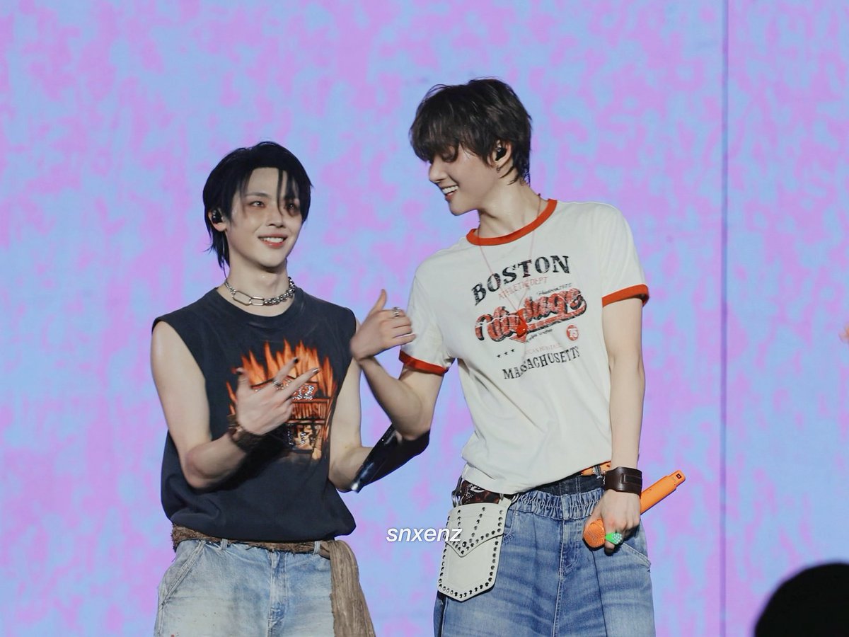 Two members of the K-pop group &TEAM share a playful moment on stage, with one wearing a black sleeveless flame-print top and chain necklace while the other sports a cream "BOSTON" ringer tee and holds an orange microphone, both smiling warmly against a pink and blue tie-dye backdrop.
