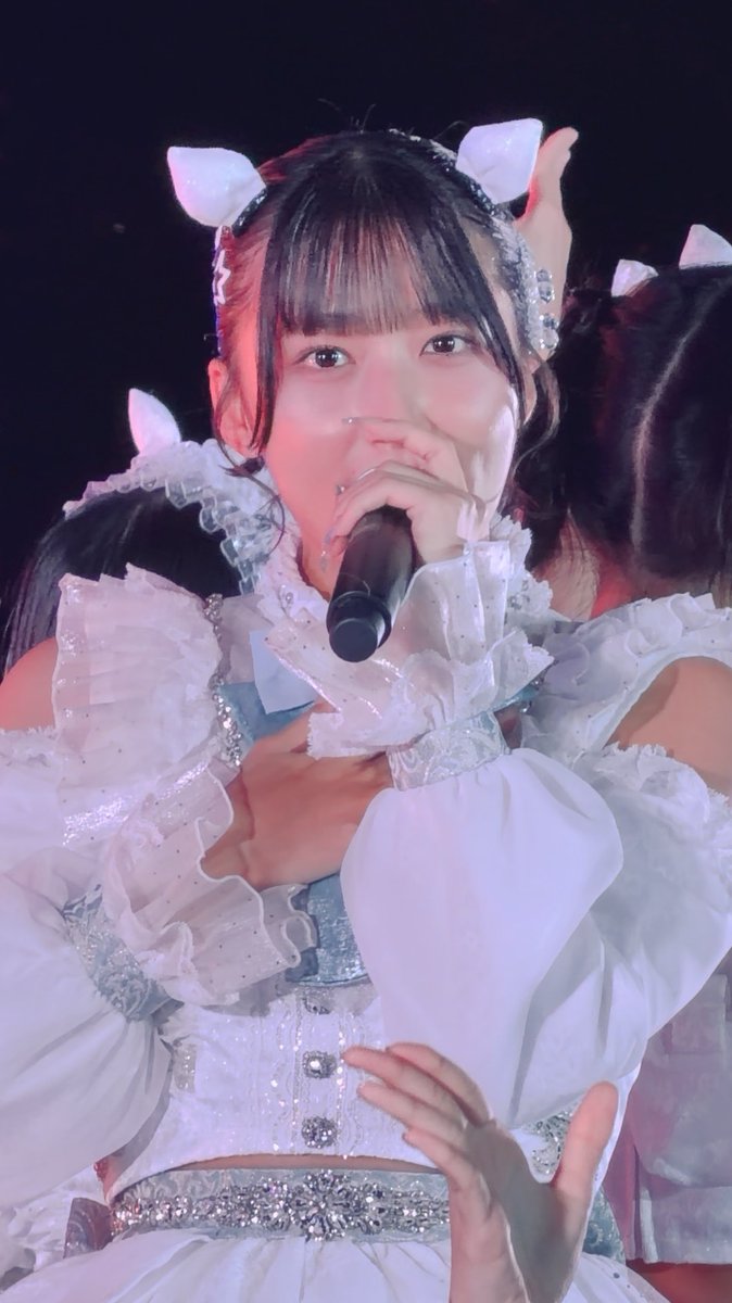 A close-up photo of a female performer on stage in a sparkly white frilled outfit with hair cat ears, holding a microphone.