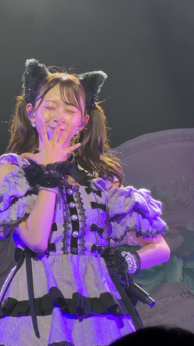 A performer with cat ear accessories and a frilly gray and black outfit sings into a microphone on stage, with eyes closed and hand raised near her face.