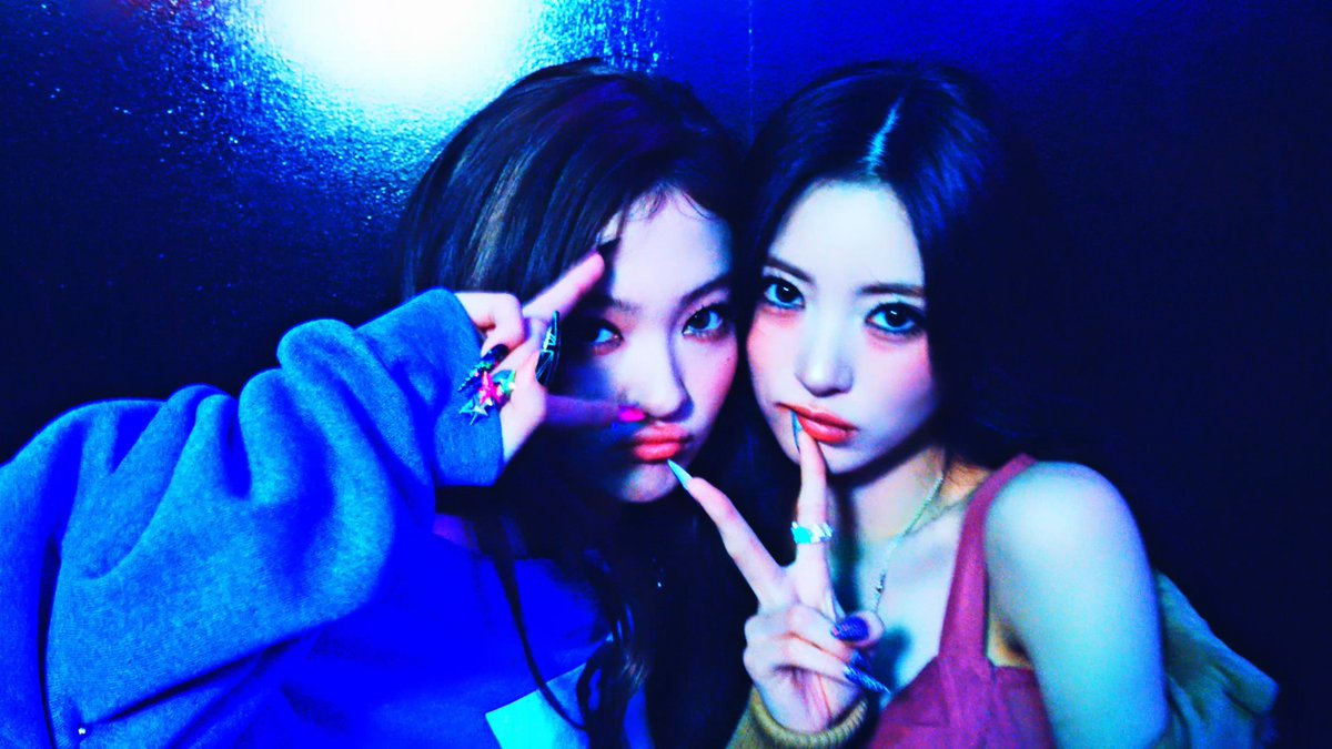 Two women pose together in blue-toned lighting, both making peace signs with their fingers near their faces.