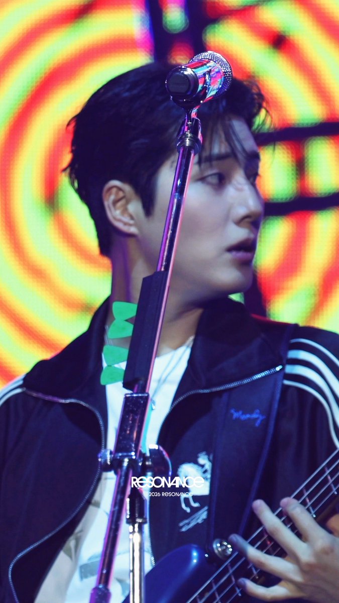 A young man with dark hair performs on stage playing a guitar and singing into a microphone, set against a vibrant background of swirling neon green, orange, and red lights. He wears a black Adidas track jacket over a white shirt, while a "2026 RESONANCE" watermark appears at the bottom of the image.