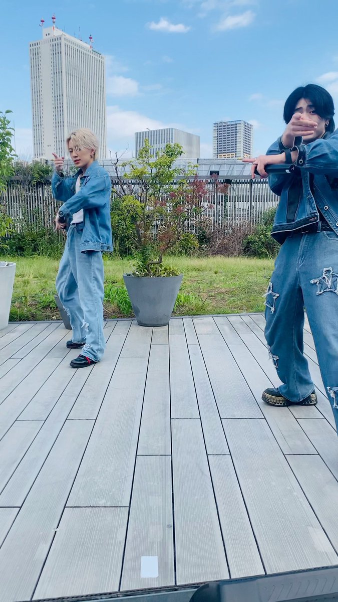 Two young men in matching denim outfits pose playfully on a wooden rooftop terrace with city skyscrapers visible in the background.