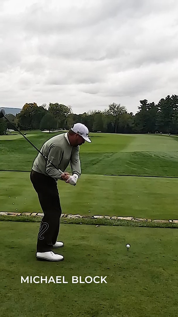 A golfer wearing a gray long-sleeve shirt, black pants, and a white cap swings his club on a tee, with the text "MICHAEL BLOCK" overlaid at the bottom. The image captures Michael Block mid-swing, highlighting the underdog story of the club pro who captivated audiences at the PGA Championship and is now competing near the top of the leaderboard on the Champions Tour.