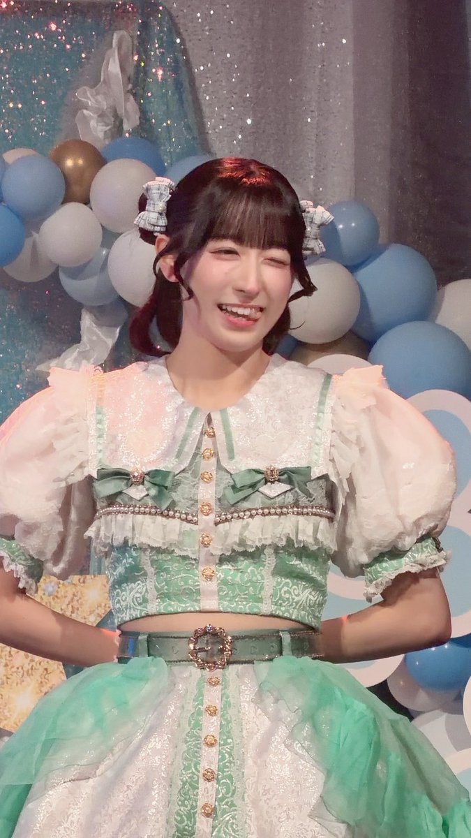 A smiling young woman in an ornate green and white frilly outfit winks at the camera, posed in front of blue and white balloons.