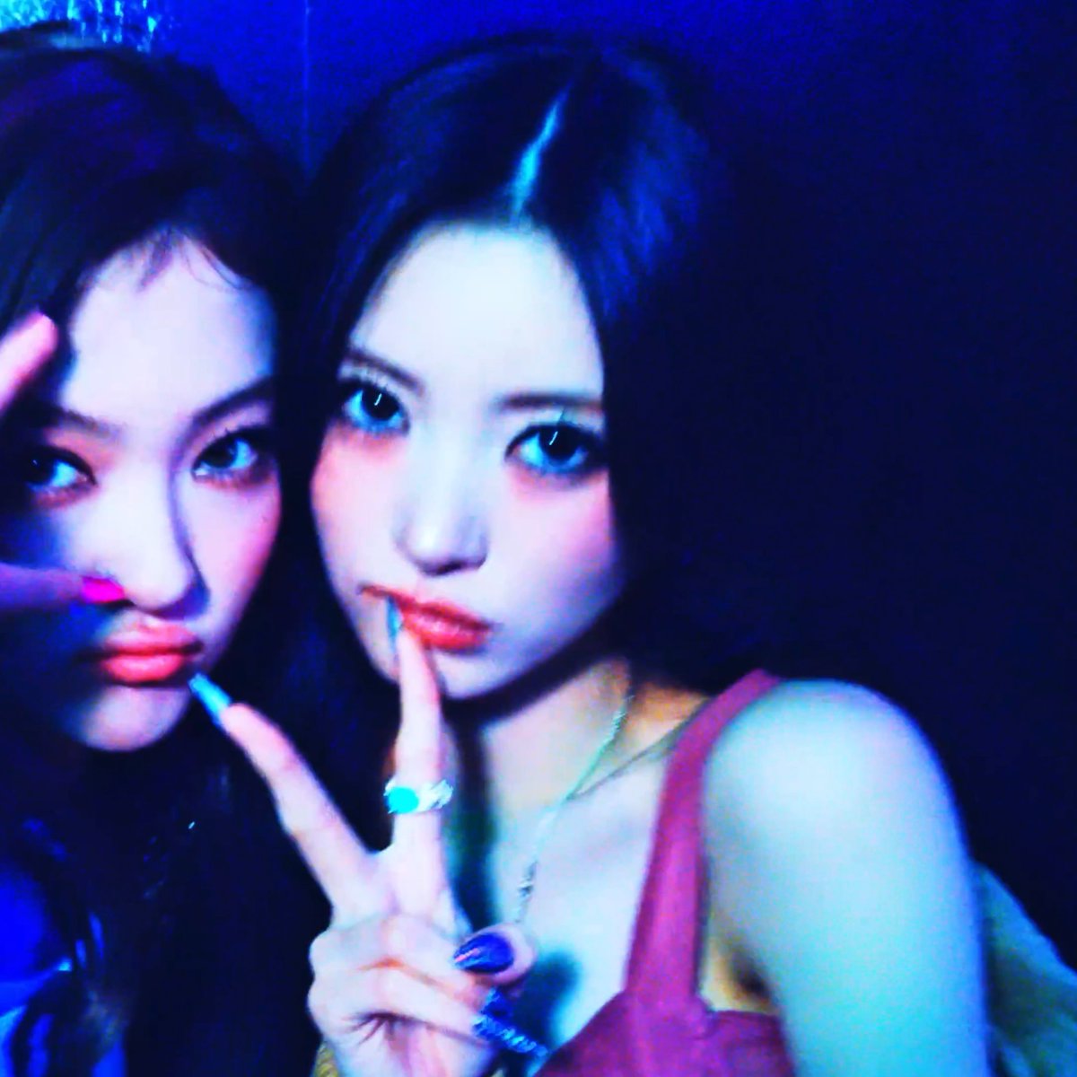 Two young women pose for a close-up photo bathed in moody blue lighting, both holding a finger to their lips in a "shh" gesture and making peace signs. The sultry, nightlife aesthetic of the image complements the bold, hyped-up caption "GO HARD ANNA," suggesting an energetic, fun-filled evening out together.