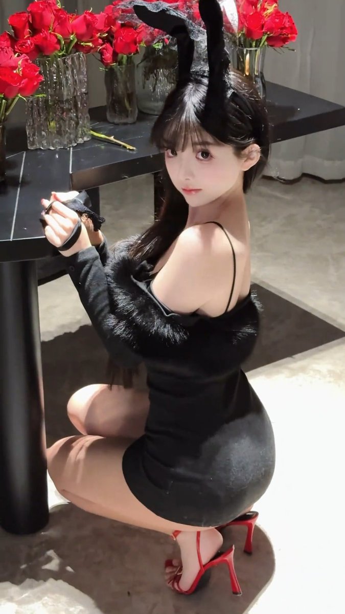 A woman wearing a black bunny ear headband and a black dress, posing with red high heels near a table of red roses.