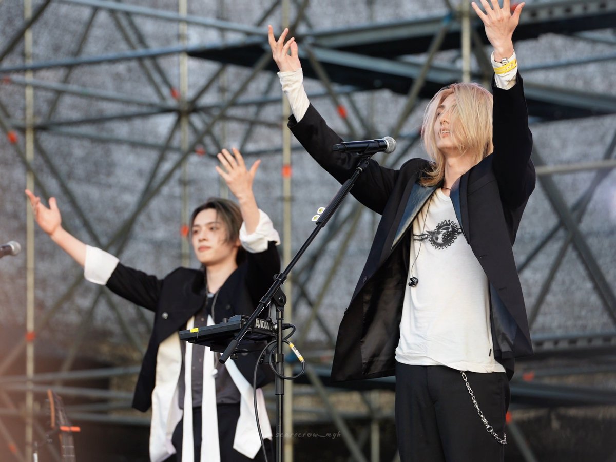 Two performers on an outdoor stage in black suits and white shirts with arms raised dramatically; one with blonde hair stands at a microphone while the other operates equipment, with 'sparecrow_mgh' watermark.