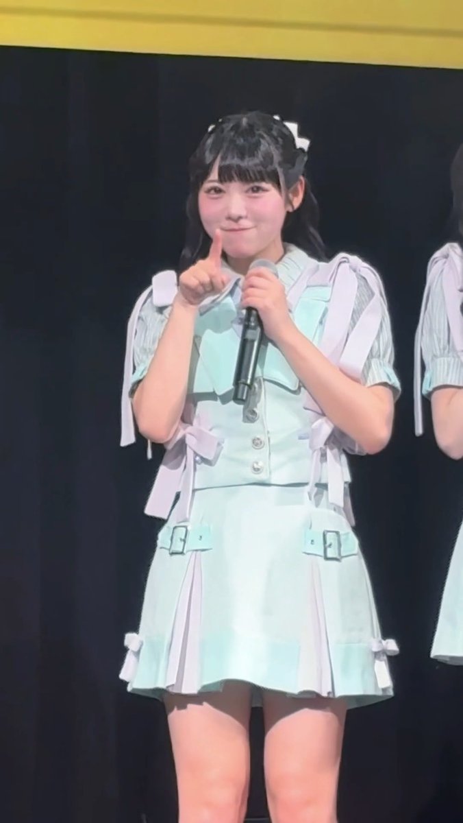 A young woman on stage wearing a mint green and white idol outfit, holding a microphone and making a peace gesture, likely a member of a Japanese idol group.