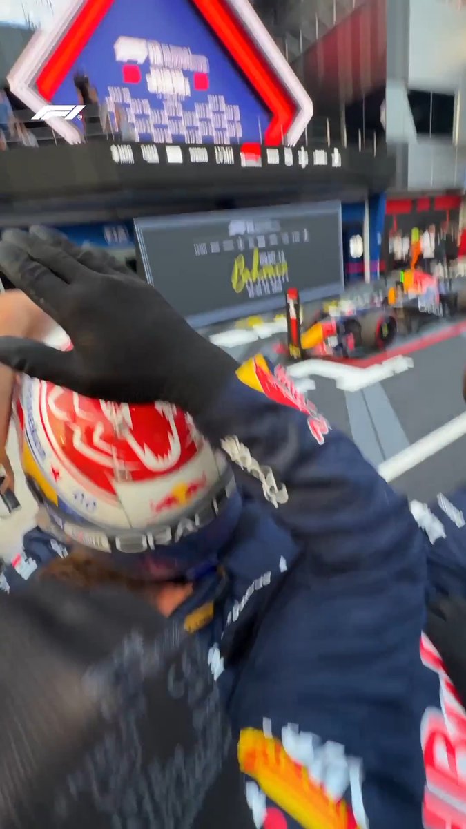 A Red Bull Racing driver, wearing a helmet featuring the iconic Red Bull logo and black gloves, celebrates in parc fermé following their victory at the Bahrain Grand Prix, with a winner's wreath visible on the F1 car in the background.