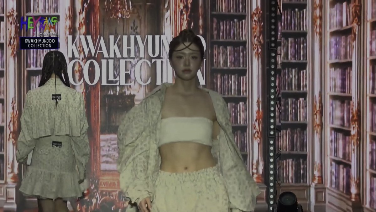 A model on a runway for the "KWAKHYUNJOO COLLECTION" wearing a light green ruffled skirt and jacket over a white bandeau top, displayed against a backdrop of a stylized library.