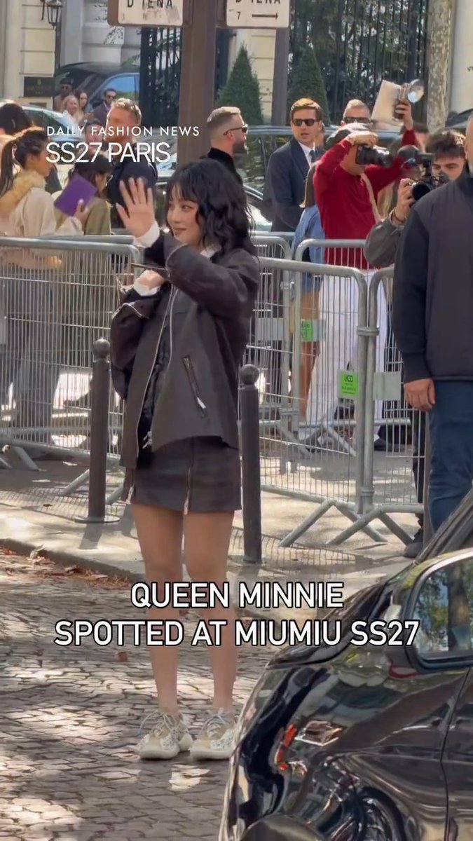A short-haired woman in a dark leather jacket and shorts waves while walking on a cobblestone street, surrounded by photographers behind a barricade, with text reading 'Daily Fashion News SS27 Paris' and 'Queen Minnie spotted at Miumiu SS27.'