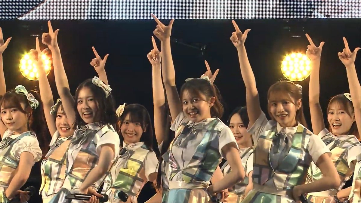 A group of young women in matching pastel plaid outfits perform on stage, raising their arms in unison with pointing fingers under bright stage lights.