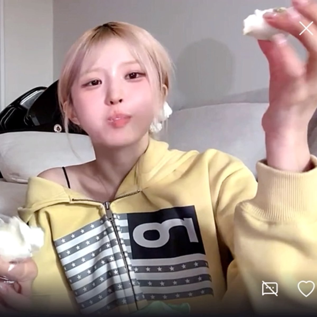 A selfie-style image of a young woman with blonde hair and bangs, wearing a yellow cropped zip-up hoodie with a graphic print, holding pieces of white food while sitting indoors.