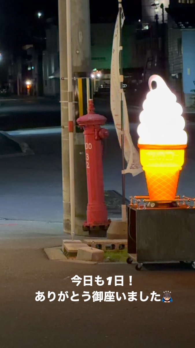 A nighttime street scene features a large illuminated ice cream cone sign next to a red Japanese fire hydrant, with Japanese text translating to 'Another day! Thank you for your patronage'.