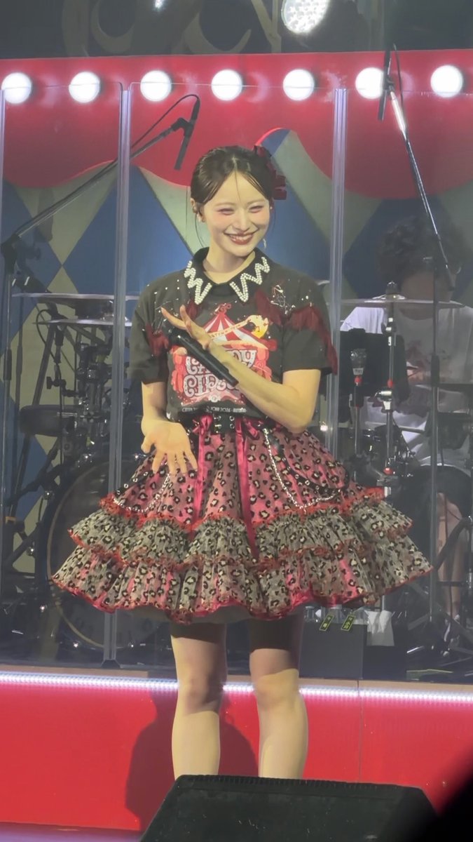 A female performer smiles on stage while wearing a black t-shirt with graphics and a detailed red and black ruffled skirt, holding a microphone.