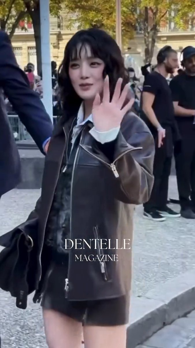A young woman with dark wavy hair and bangs waving at the camera while wearing a brown leather jacket over a dark outfit, with 'DENTELLE MAGAZINE' watermarked across the image.