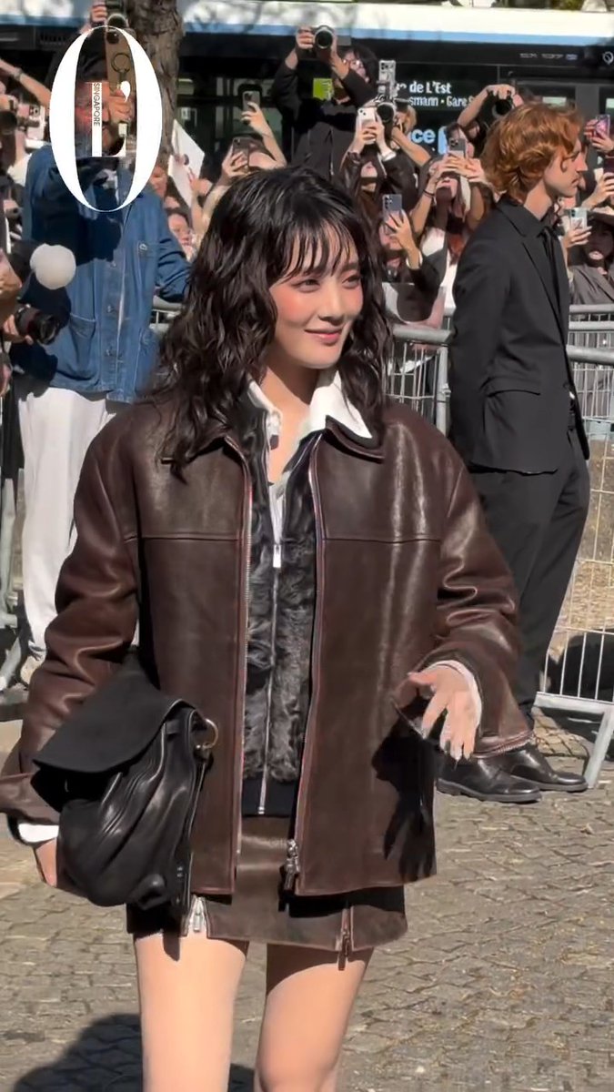 A young woman with wavy black hair wearing a brown leather jacket walks past a crowd of photographers and fans, with one person holding a camera circled in white.