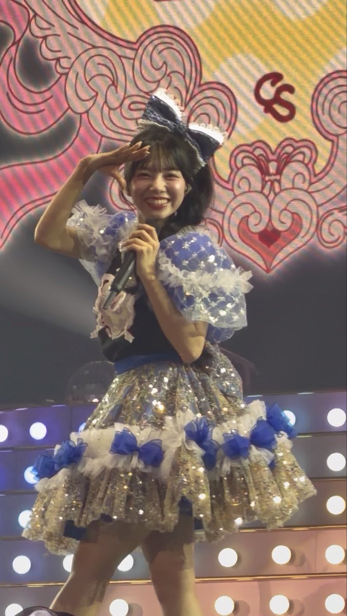 A performer in a blue and silver sequined outfit with white lace details smiles and salutes while holding a microphone on a stage with an ornate backdrop.