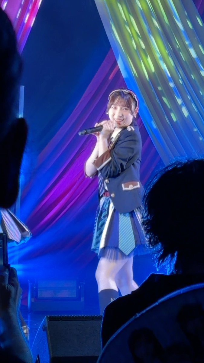 A young woman performing on stage, holding a microphone and smiling, wearing a blue blazer with decorative elements and a pleated skirt against a blue and purple striped backdrop.
