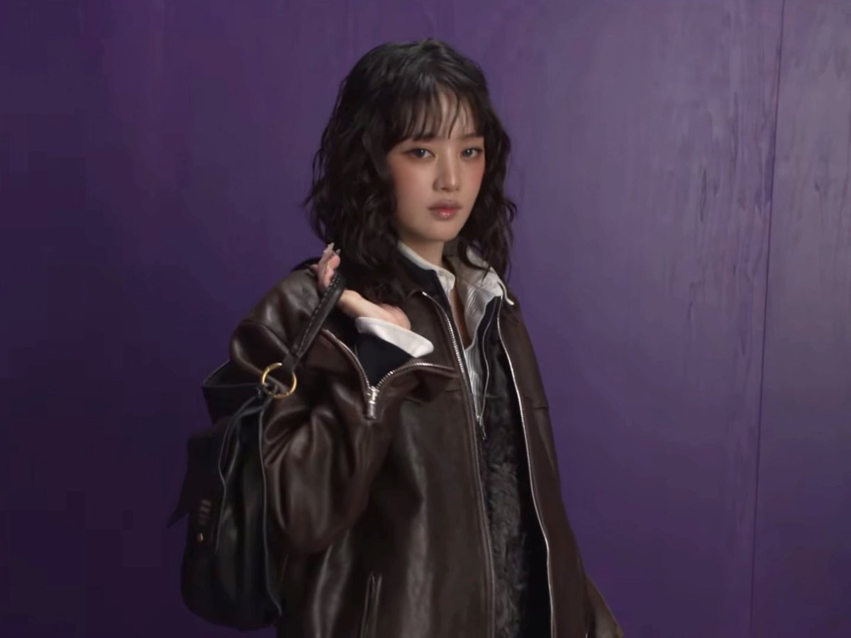 A portrait of a young woman with short dark hair, wearing a brown leather jacket over a white shirt, posing against a purple background.
