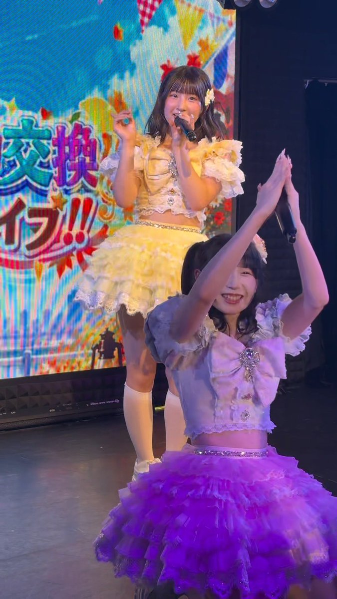 Two young women in frilly yellow and purple costumes perform on stage, with one singing into a microphone in front of a colorful screen displaying Japanese text including '交換ライブ!!'.