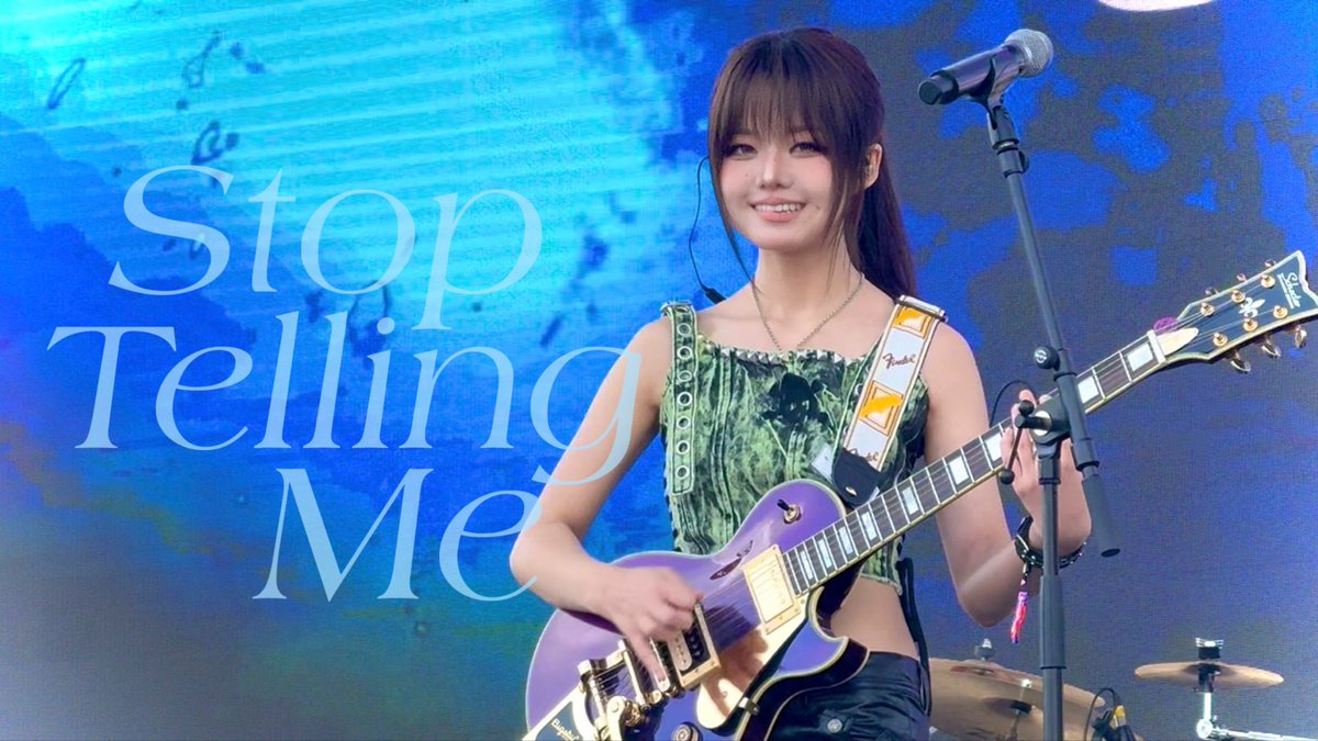 A woman performs on stage playing a purple electric guitar (Fender Les Paul) while smiling, with a blue backdrop displaying the text 'Stop Telling Me'.