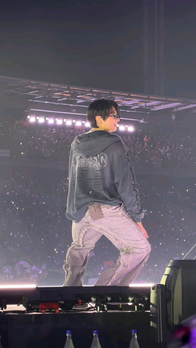 A performer stands on stage in a gray hoodie reading "ADDICTION" and distressed jeans, looking over his shoulder toward a sea of fans holding up light sticks during a concert.
