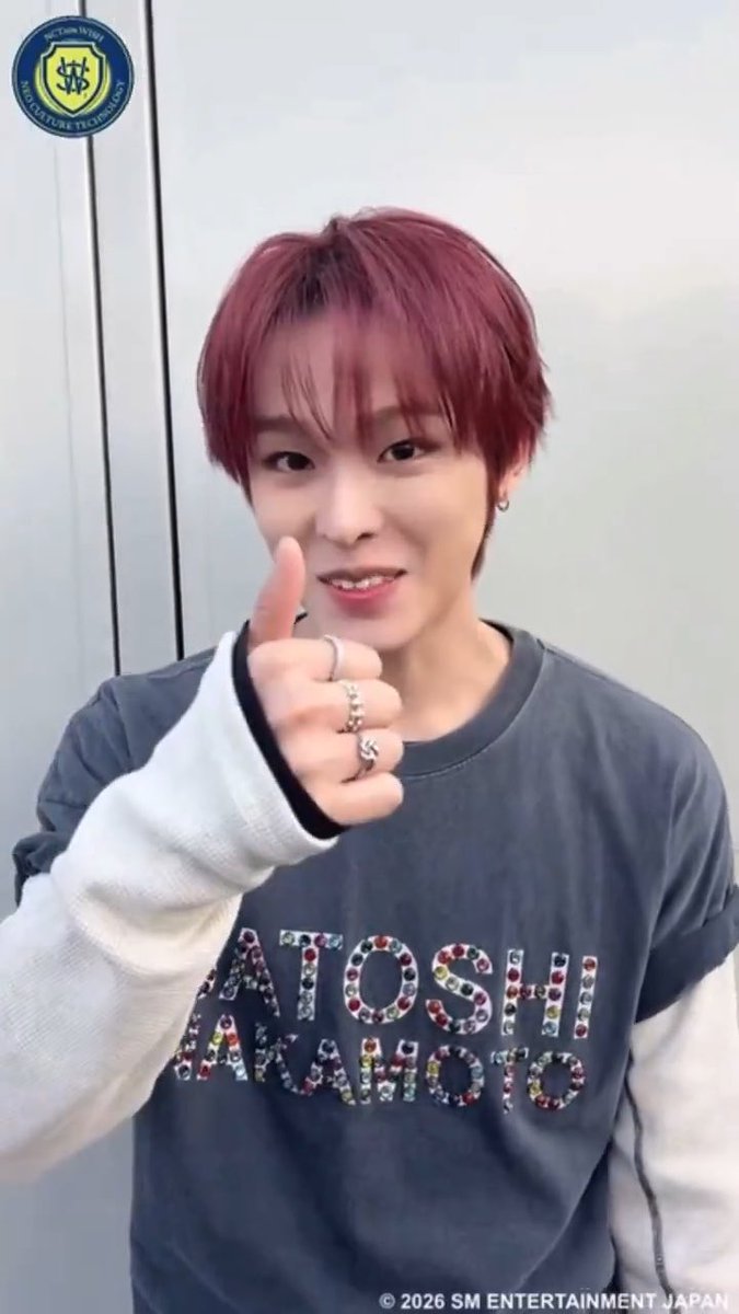A selfie-style image of a smiling young man with pink hair giving a thumbs up, wearing a grey 'ATOSHI WAKAOTO' t-shirt over white long sleeves, with rings on his fingers and the SM Entertainment Japan copyright.