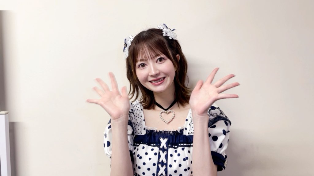 A smiling young woman with shoulder-length brown hair and bangs wears a polka-dot dress with a heart pendant necklace, waving both hands at the camera.
