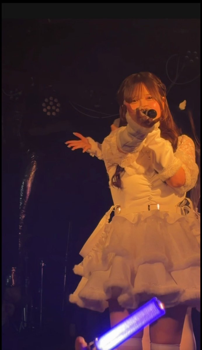 A woman in a frilly white outfit performs on stage at a concert, holding a microphone and pointing toward the audience.