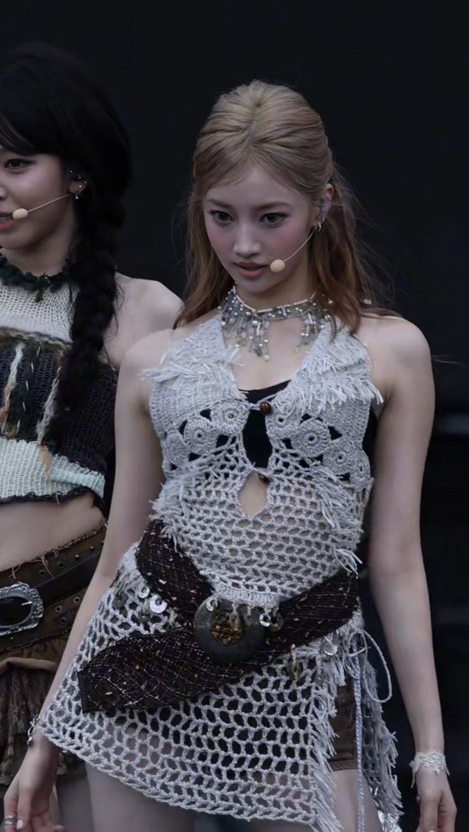 A photograph of K-pop idol Ningning from aespa performing on stage, wearing a white crocheted halter top with fringe details and a large belt buckle, alongside another member.