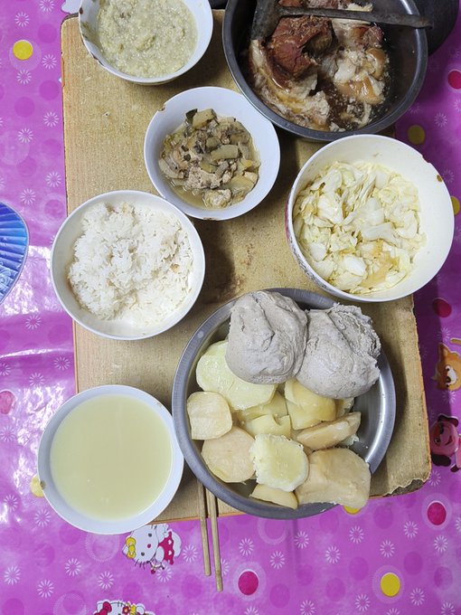A top-down view of a traditional meal laid out on a wooden board with multiple bowls containing rice, soup, vegetables, mushrooms, steamed buns, and sliced taro or sweet potato, alongside a Hello Kitty themed placemat.