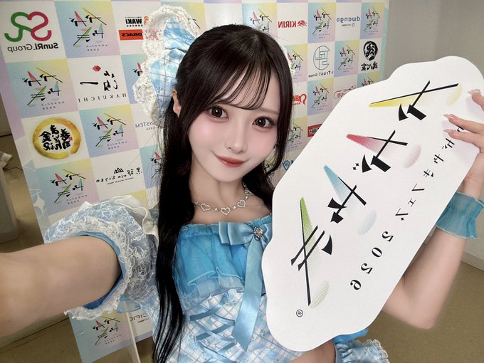 A woman with long dark hair, wearing a blue and white outfit, holds a paddle displaying Japanese text while posing in front of a backdrop covered in logos.
