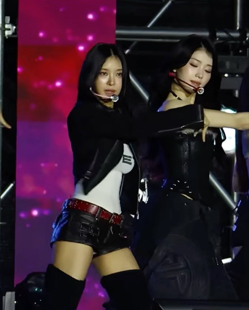 Two female K-pop performers dance on stage in edgy black outfits with leather shorts and boots against a pink and dark background.