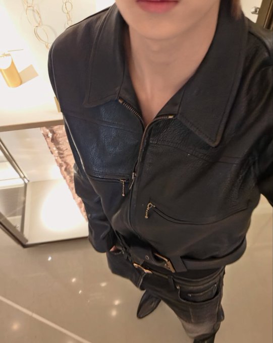 A person models a black leather jacket and matching leather pants, with the camera capturing them from above in what appears to be a luxury boutique setting. The image humorously highlights both the leather jacket's front zipper details and the coordinating bottoms, matching the post's playful comment about capturing both pieces in separate shots.