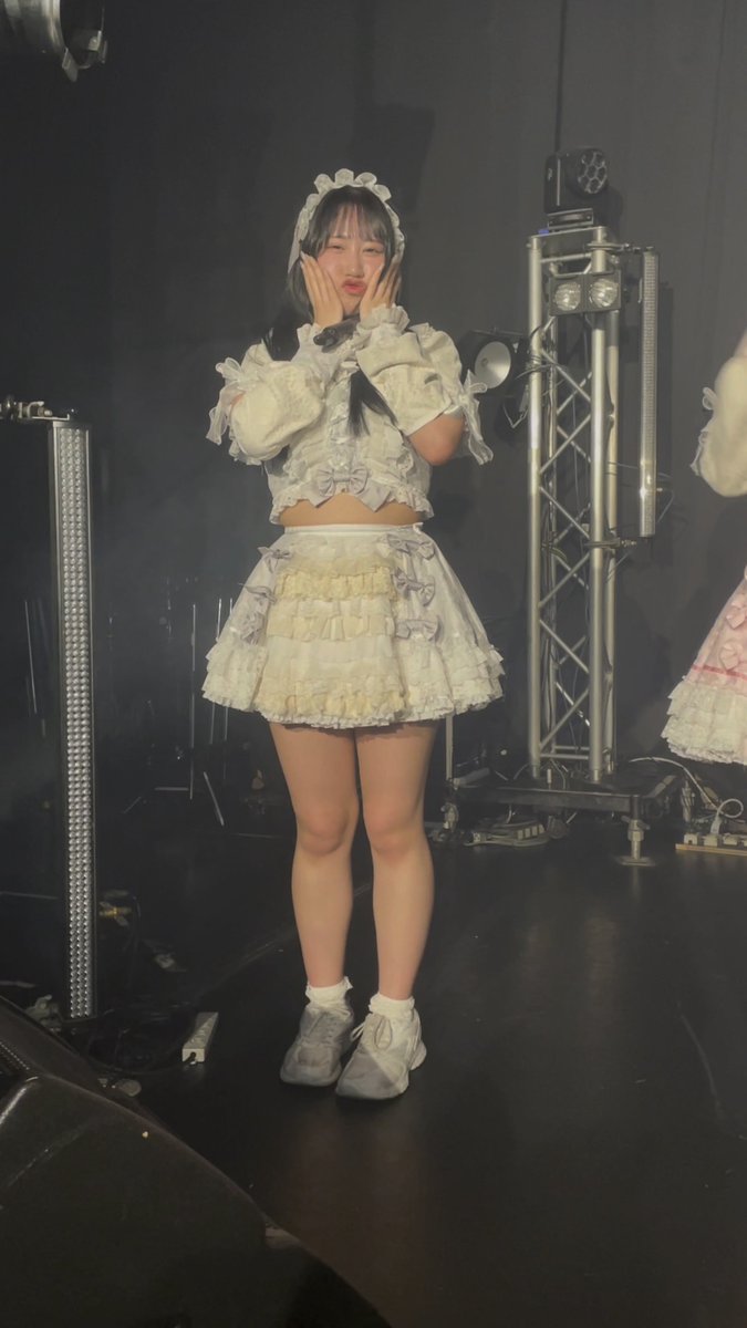 A female performer in a frilly white and gray outfit with a headband poses with her hands on her cheeks on a dimly lit stage with lighting equipment.