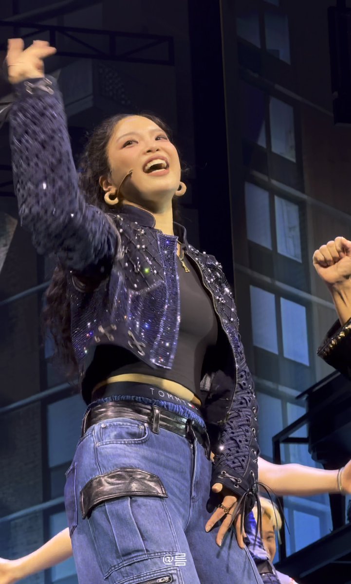 A female performer on stage wearing a sequined purple jacket and Tommy Hilfiger jeans, raising her arm while smiling.