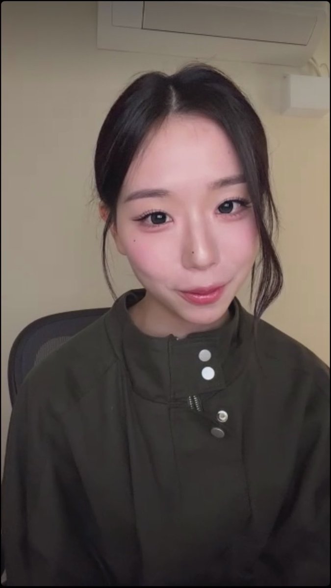 A close-up portrait of a young woman with dark hair pulled back, wearing a dark green jacket, looking directly at the camera.