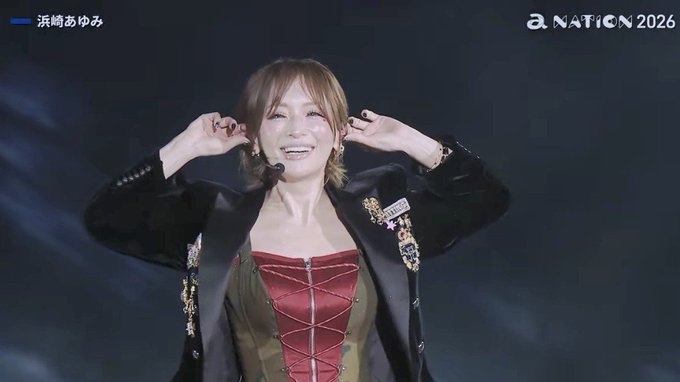 浜崎あゆみ smiles brightly while performing at "a NATION 2026," adjusting her earrings near a headset microphone while dressed in a red corset top with a black jacket.