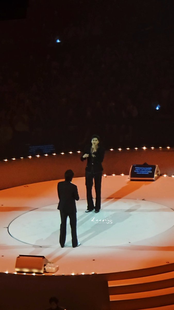 Two performers stand on a circular stage lit with candles during what appears to be a theatrical or musical performance.