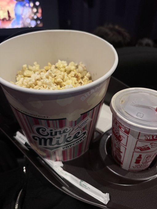 A large Cine Mike popcorn tub and a branded drink cup rest on a tray in a darkened room, likely a theater.
